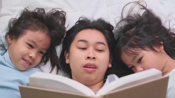 Υπέροχη ευτυχισμένη ασιατική οικογένεια στο άνετο σπίτι. Όμορφη νεαρή μητέρα διαβάζει ένα βιβλίο στις κόρες της σε ένα κρεβάτι στο σπίτι. Περνάμε καλά στο σπίτι.. - Πλάνα, βίντεο