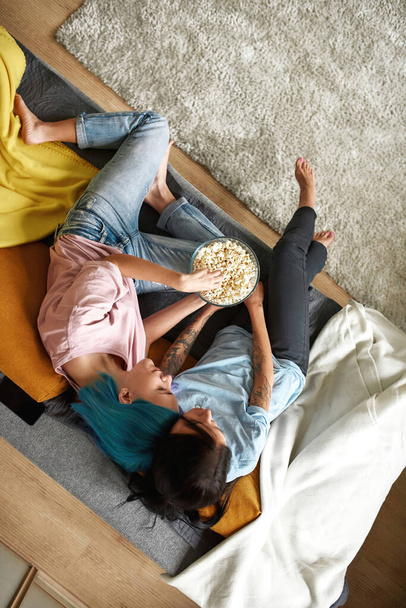 Lesbian couple watching TV on sofa, eating popcorn - Фото, изображение