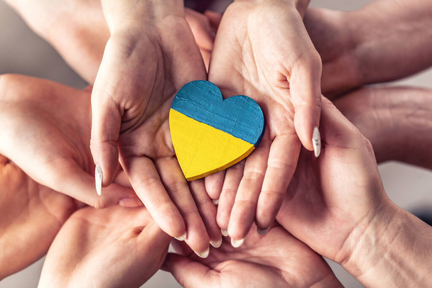 Wir stehen mit dem ukrainischen Symbol, während zahlreiche Hände sich in Einheit halten, mit einem hölzernen Herz, das in den Farben der ukrainischen Flagge bemalt ist.. - Foto, Bild