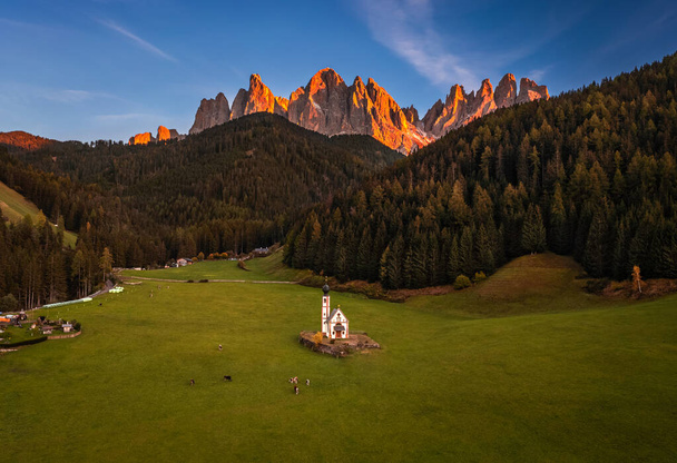 Val Di Funes, Dolomity, Włochy - Widok z lotu ptaka na piękny kościół św. Jana (Chiesetta di San Giovanni in Ranui) w południowym Tyrolu z włoskimi Dolomitami w ciepłych kolorach zachodu słońca na tle  - Zdjęcie, obraz