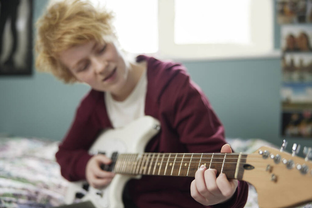 Nastolatka siedzi na łóżku ucząc się grać na gitarze elektrycznej w sypialni - Zdjęcie, obraz