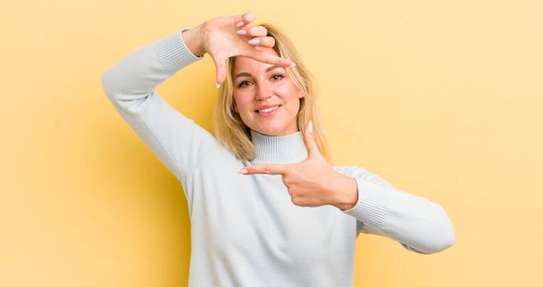 blonde caucasian woman feeling happy, friendly and positive, smiling and making a portrait or photo frame with hands - 写真・画像