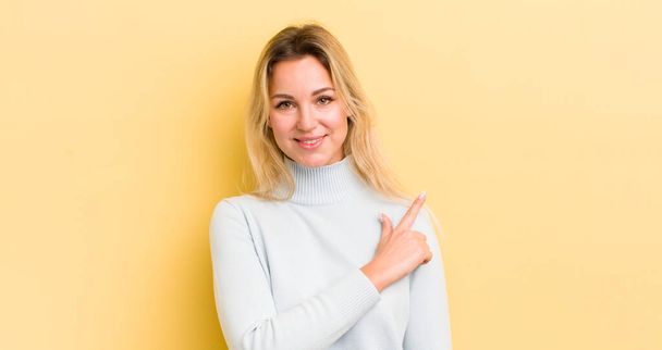 blonde caucasian woman smiling cheerfully, feeling happy and pointing to the side and upwards, showing object in copy space - Fotoğraf, Görsel