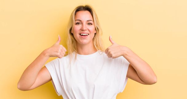 blond Kaukasische vrouw glimlachen breed op zoek gelukkig, positief, zelfverzekerd en succesvol, met beide duimen omhoog - Foto, afbeelding
