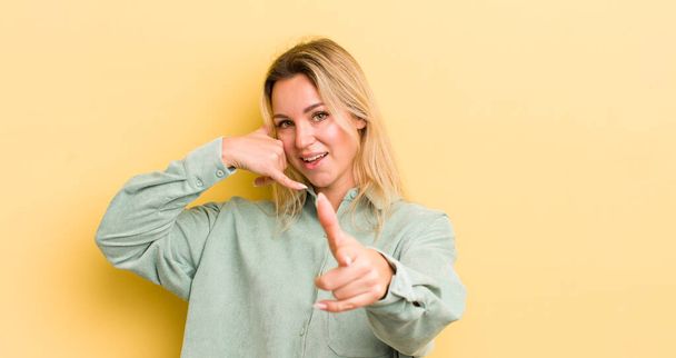 blonde caucasian woman smiling cheerfully and pointing to camera while making a call you later gesture, talking on phone - Fotó, kép