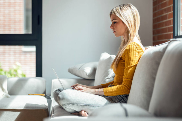 Tiro de bela jovem mulher trabalhando com seu laptop enquanto sentado em um sofá em casa. - Foto, Imagem