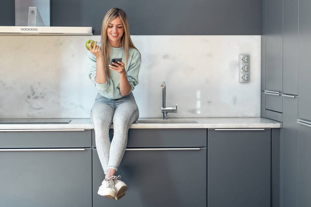 Shot of pretty young woman using her mobile phone while eating an apple sitting on the kitchen at home. - Foto, imagen