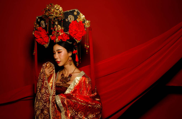 Dramatic Series femenino expresa sentirse emocional con la tela revoloteando en el aire. Mujer asiática usan traje tradicional de la emperatriz real de China con vestido de diseño de línea de oro y ropa de cabeza, fondo rojo - Foto, imagen