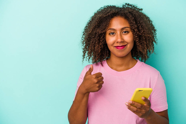 若いアフリカ系アメリカ人女性の携帯電話を保持青の背景笑顔と親指を上げる - 写真・画像