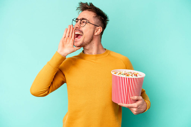 młody biały mężczyzna jedzenie popcorn odizolowany na niebieskim tle krzycząc i trzymając dłoń blisko otwarte usta. - Zdjęcie, obraz