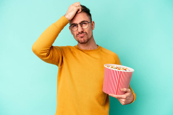 Junge kaukasische Mann essen Popcorn isoliert auf blauem Hintergrund schockiert, sie erinnert sich an wichtiges Treffen. - Foto, Bild