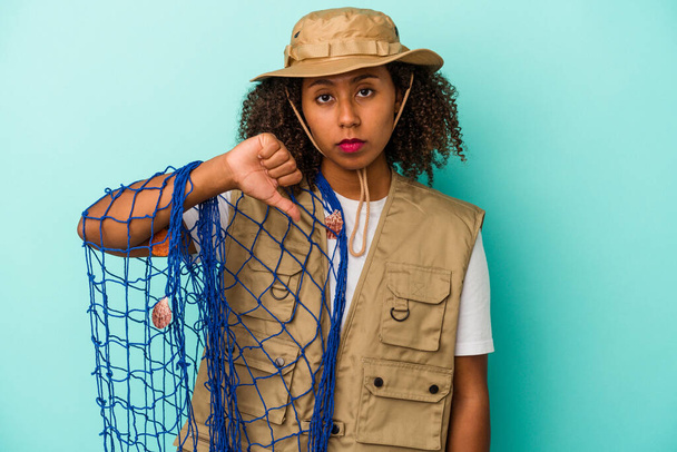 Young african american fisherwoman holding net isolated on blue background  showing a dislike gesture, thumbs down. Disagreement concept. - Photo, Image
