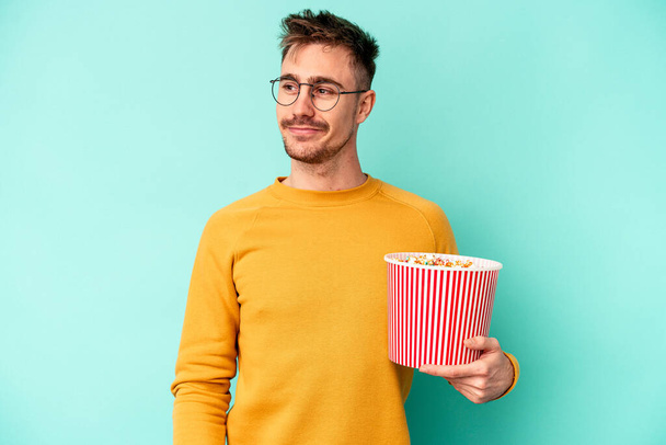 Junger kaukasischer Mann, der Popcorn isoliert auf blauem Hintergrund isst, blickt lächelnd, fröhlich und angenehm zur Seite. - Foto, Bild