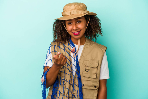 Fiatal afro-amerikai halász nő tartja háló elszigetelt kék háttér mutató ujjal rád, mintha hívogató közelebb jön. - Fotó, kép