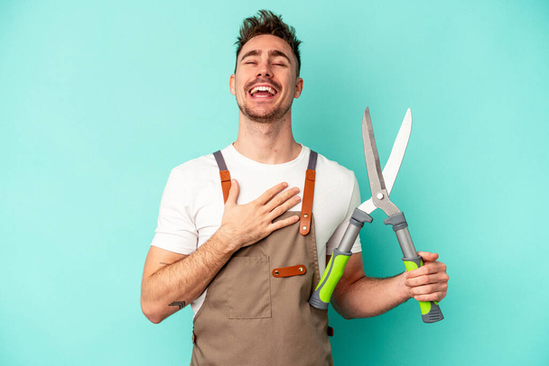 Young gardener caucasian man holding a scissors isolated on blue background laughs out loudly keeping hand on chest. - Photo, Image