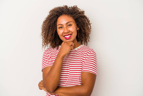 jong Afrikaans amerikaans vrouw met krullend haar geïsoleerd op witte achtergrond glimlachen gelukkig en zelfverzekerd, aanraken kin met de hand. - Foto, afbeelding