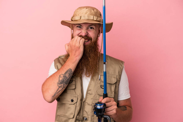 Joven pescador de jengibre caucásico con barba larga sosteniendo una caña aislada sobre fondo rosa mordiendo las uñas, nervioso y muy ansioso. - Foto, Imagen