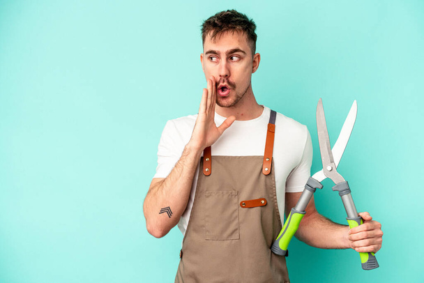 Young gardener caucasian man holding a scissors isolated on blue background is saying a secret hot braking news and looking aside - Photo, Image