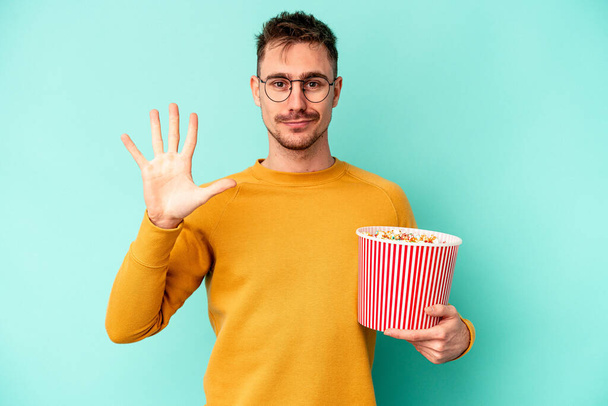 Junger kaukasischer Mann isst Popcorn isoliert auf blauem Hintergrund und lächelt fröhlich und zeigt Nummer fünf mit den Fingern. - Foto, Bild