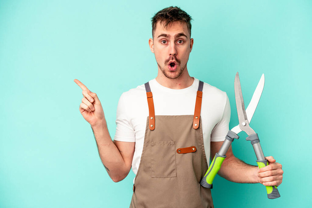 Young gardener caucasian man holding a scissors isolated on blue background pointing to the side - Photo, Image
