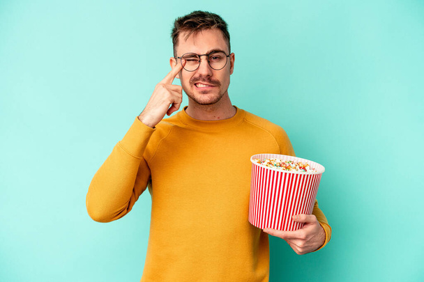 Junger kaukasischer Mann isst Popcorn isoliert auf blauem Hintergrund und zeigt eine enttäuschende Geste mit dem Zeigefinger. - Foto, Bild