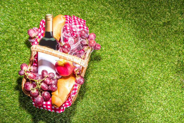 Picknickmand met eten, wijnfles, glazen en fruit op groen zonnig gazongras  - Foto, afbeelding