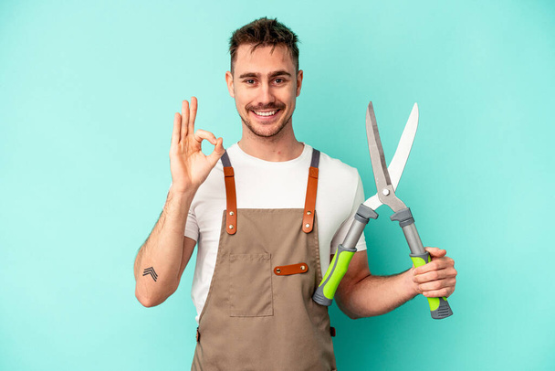 Young gardener caucasian man holding a scissors isolated on blue background cheerful and confident showing ok gesture. - Photo, Image