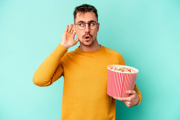 Junger kaukasischer Mann isst Popcorn isoliert auf blauem Hintergrund und versucht Klatsch und Tratsch zu hören. - Foto, Bild