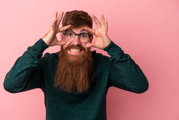 Молодой кавказский рыжий мужчина с длинной бородой, изолированный на розовом фоне, держит глаза открытыми, чтобы найти возможность для успеха. - Фото, изображение