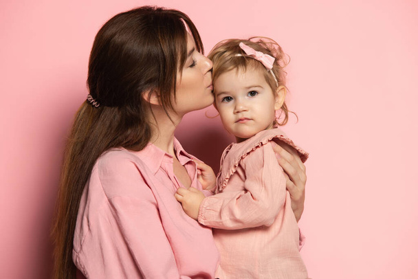 Kiss. Portrait of young woman and little girl, mother and daughter isolated on pink studio background. Mothers Day celebration. Concept of family, childhood, motherhood - Foto, Bild