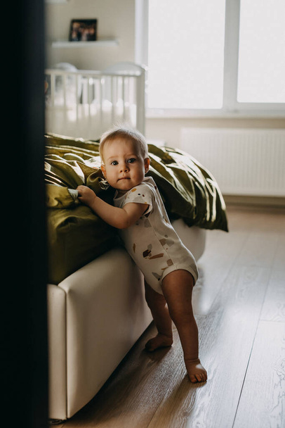 Baby staat naast een bed, leunend erop, blootsvoets, thuis, kijkend naar de camera. - Foto, afbeelding