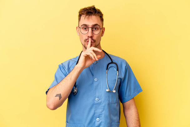 Νεαρός νοσοκόμος καυκάσιος άνδρας απομονωμένος σε κίτρινο φόντο κρατώντας μυστικό ή ζητώντας σιωπή. - Φωτογραφία, εικόνα