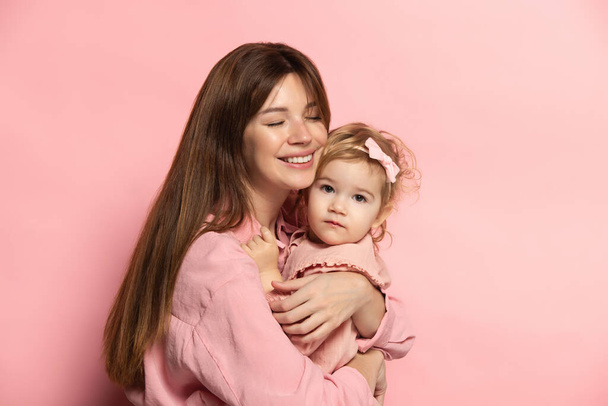 Huggins. Portrait of young woman and little girl, mother and daughter isolated on pink studio background. Mothers Day celebration. Concept of family, childhood, motherhood - Foto, Imagen