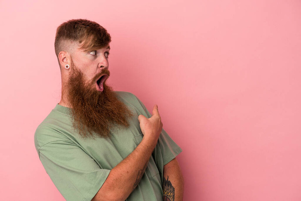 Giovane uomo zenzero caucasico con lunga barba isolata su punti di fondo rosa con il pollice lontano, ridendo e spensierato. - Foto, immagini
