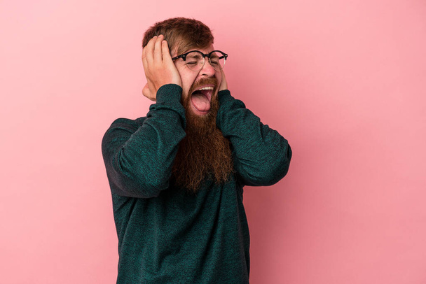 Jovem homem de gengibre caucasiano com barba longa isolado no fundo rosa cobrindo orelhas com as mãos tentando não ouvir som muito alto. - Foto, Imagem