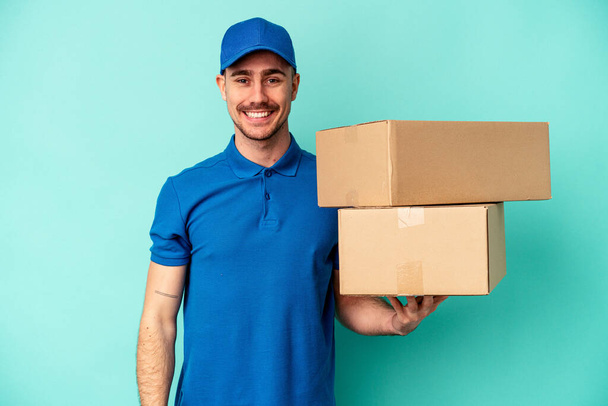 Young delivery caucasian man isolated on blue background happy, smiling and cheerful. - 写真・画像