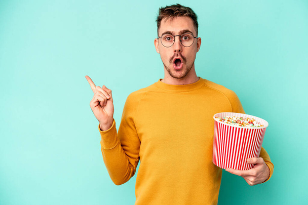 Young caucasian man eating popcorn isolated on blue background pointing to the side - Fotoğraf, Görsel
