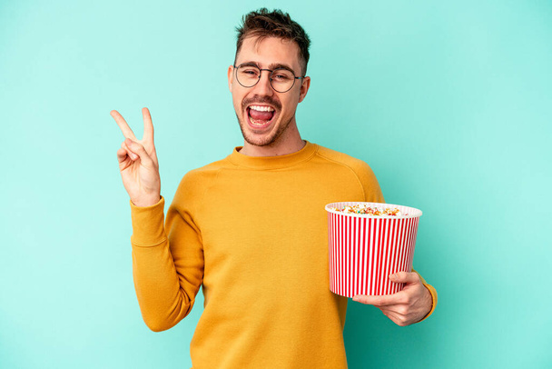 Junger kaukasischer Mann isst Popcorn isoliert auf blauem Hintergrund und zeigt fröhlich und unbeschwert ein Friedenssymbol mit Fingern. - Foto, Bild