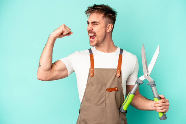 Young gardener caucasian man holding a scissors isolated on blue background raising fist after a victory, winner concept. - Photo, Image