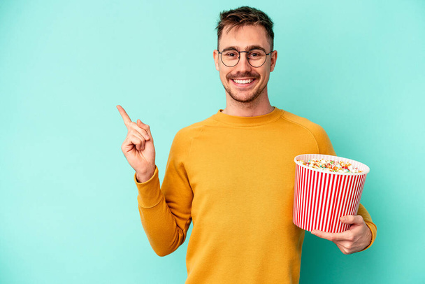 Fiatal kaukázusi férfi eszik popcorn elszigetelt kék háttér mosolyog és rámutatva félre, mutat valamit az üres térben. - Fotó, kép