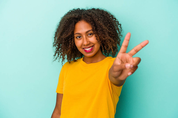 Junge afrikanisch-amerikanische Frau mit lockigem Haar isoliert auf blauem Hintergrund fröhlich und unbeschwert zeigt ein Friedenssymbol mit Fingern. - Foto, Bild