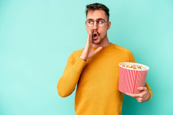 Giovane uomo caucasico mangiare popcorn isolato su sfondo blu sta dicendo una notizia segreta frenante caldo e guardando da parte - Foto, immagini