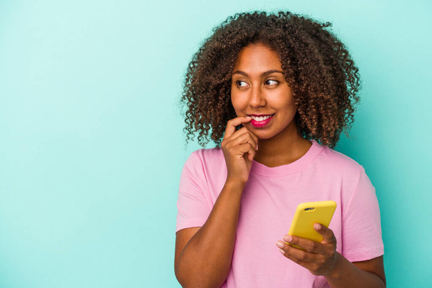 Jeune femme afro-américaine tenant un téléphone portable isolé sur fond bleu détendu penser à quelque chose en regardant un espace de copie. - Photo, image