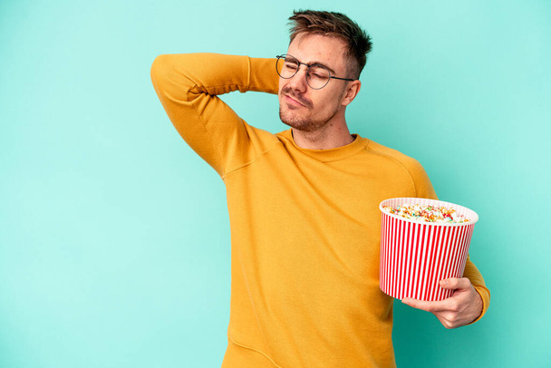 Nuori valkoihoinen mies syö sinisellä taustalla eristettyä popcornia, koskettaa pään takaosaa, ajattelee ja tekee valinnan.. - Valokuva, kuva