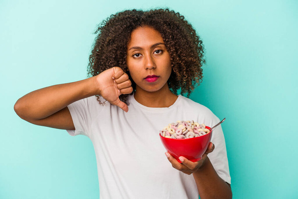 Young african american woman holding a bowl of cereals isolated on blue background showing a dislike gesture, thumbs down. Disagreement concept. - Photo, Image