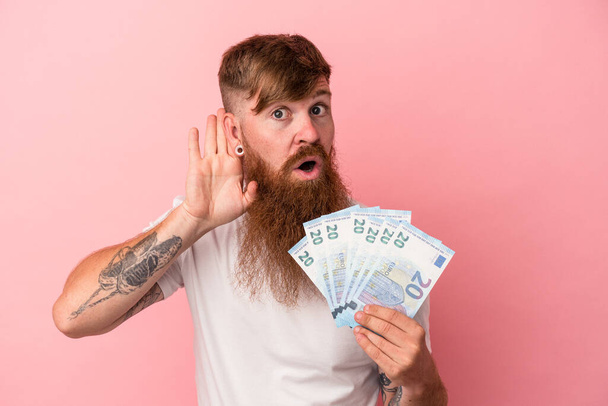 Young caucasian ginger man with long beard holding banknotes isolated on pink background trying to listening a gossip. - Fotografie, Obrázek