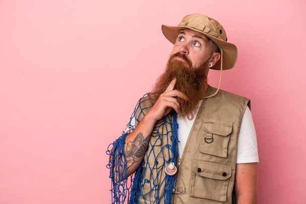 Young caucasian ginger fisherman with long beard holding a net isolated on pink background - 写真・画像