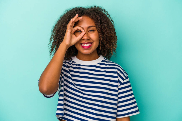 Młoda Afroamerykanka z kręconymi włosami odizolowana na niebieskim tle podekscytowana trzymaniem ok gestu na oku. - Zdjęcie, obraz