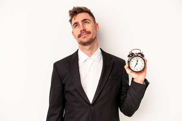 Young caucasian business man holding alarm clock isolated on white background dreaming of achieving goals and purposes - Fotó, kép
