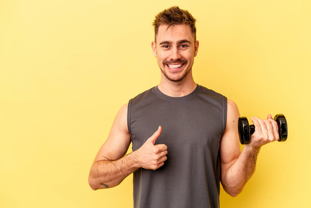 Молодий спортивний чоловік тримає гантелі ізольовані на жовтому тлі посміхаючись і піднімаючи великий палець вгору
 - Фото, зображення